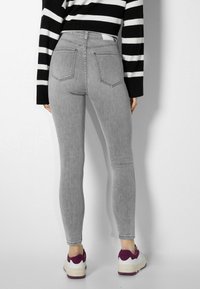 Jeans ajustados de tiro alto en gris, con textura desgastada, diseño de cinco bolsillos, combinados con zapatillas deportivas blancas y moradas, mostrados desde la parte trasera.
