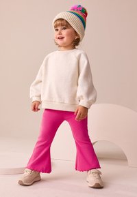 Ljusbeige fleece-tröja med ballongärmar, kombinerad med knallrosa utsvängda byxor och beiga sneakers, kompletterat med en flervägs pom-pom mössa.
