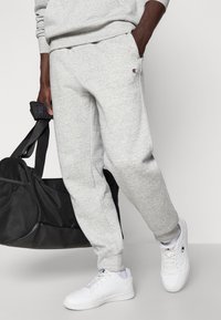 Γκρι φόρμες από βαμβακερό μείγμα, με ελαστική μέση, πλαϊνές τσέπες και λεπτομέρεια λογότυπου. Συνδυάζονται με λευκά αθλητικά παπούτσια και μαύρη τσάντα.