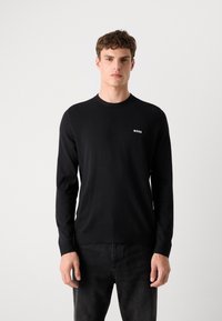 Schwarzer Pullover mit langen Ärmeln aus glattem Strick und Rundhalsausschnitt. Kleines weißes "BOSS"-Logo auf der Brust. Kombiniert mit dunkler Jeans.
