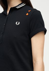Femme portant un polo noir avec un logo de laurier blanc sur la poitrine et deux petites patchs de cœur orange sur l'épaule.