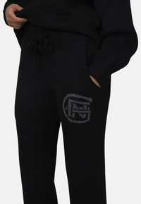 Czarne dzianinowe spodnie dresowe z elastycznym paskiem na sznurki, bocznymi kieszeniami i szarym logo naszytym na lewej udzie. Miękka tekstura, luźny krój.