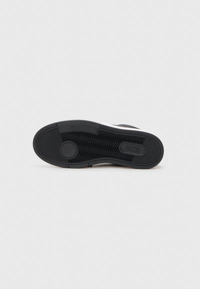 Suela de zapatilla negra de goma con un patrón texturizado de espiga; presenta un emblema circular cerca del talón y acentos laterales lisos.