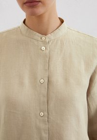Beige linen shirt with a mandarin collar, featuring four small round buttons down the front. Soft texture and simple design.