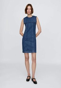 Denim ermeløs kjole med firkantet halslinning, vertikale sømmer, og rett snitt. Mørkeblå farge og lett tekstur. Sorte flate sko.