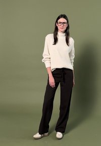 Femme aux longs cheveux foncés portant des lunettes, un pull en maille crème, un pantalon noir et des chaussures blanches, debout devant un fond vert olive.