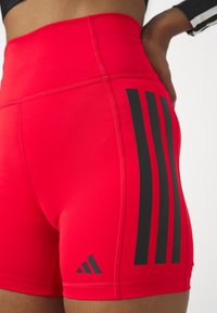 Crvene atletske kratke hlače od rastezljivog materijala, s visokim pojasom i tri crne vertikalne pruge sa strane, s Adidas simbolom.
