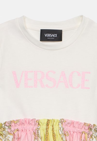 Ρούχο σε κρεμ χρώμα με ροζ γκλίτερ λογότυπο Versace στο στήθος και ύφασμα με πολύχρωμο σχέδιο σε ροζ, κίτρινο και χρυσό από κάτω.