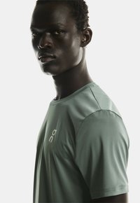 Tânăr bărbat de culoare cu părul scurt, purtând un tricou sportiv de culoare verde deschis, privind direct în față cu o expresie neutră.