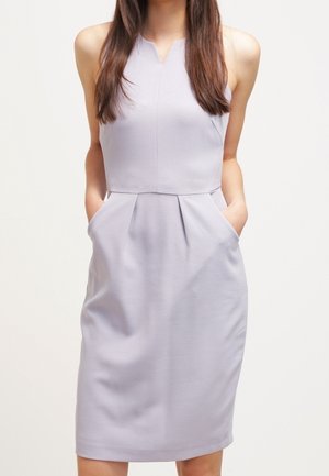 Robe sans manches gris clair, longueur genou, avec poches avant, taille cintrée et léger décolleté en V, portée par une personne aux longs cheveux.