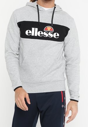 Mężczyzna ubrany w jasnoszarą bluzę z kapturem z czarną wstawką na klatce piersiowej i białym logo "ellesse", w połączeniu z granatowymi spodniami ozdobionymi paskami z logo po bokach.