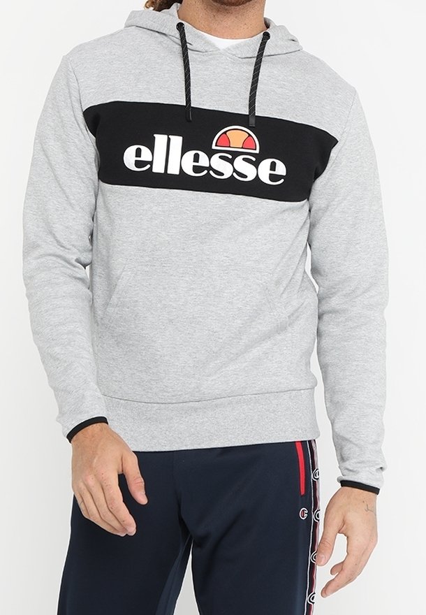 Homme portant un sweat à capuche gris clair avec un panneau noir sur la poitrine et un logo blanc "ellesse", associé à un pantalon bleu marine avec des bandes de logo sur le côté.