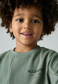 Sudadera verde con acabado texturizado, con un cuello redondo y logo impreso en negro. Mangas cortas y detalles de costura visibles.