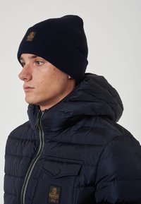 Cappello a maglia blu navy e giacca imbottita con texture trapuntata, dotata di tasca sul petto e patch con logo sulla giacca.