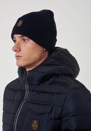Cappello a maglia blu navy e giacca imbottita con texture trapuntata, dotata di tasca sul petto e patch con logo sulla giacca.
