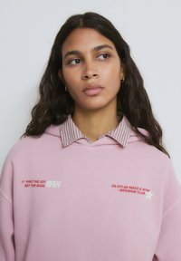 Sudadera rosa de gran tamaño con texto bordado en blanco y rojo, superpuesta sobre una camisa con cuello y patrón a rayas rojas.