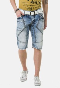 Denim-Shorts in Hellblau mit einem ausgewaschenen Look, der einzigartige gestickte Muster, Vordertaschen und einen weißen Gürtel umfasst. Die Schuhe sind weiße Sneakers.