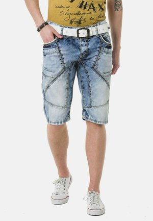 Denim-Shorts in Hellblau mit einem ausgewaschenen Look, der einzigartige gestickte Muster, Vordertaschen und einen weißen Gürtel umfasst. Die Schuhe sind weiße Sneakers.