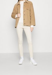 Ljusbrun ulljacka med krage och knappar fram, kombinerad med en krämfärgad kabelstickad tröja och krämfärgade skinny-byxor, avslutad med vita sneakers.