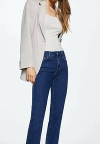 Blazer beige sobre una camiseta sin mangas clara, combinado con jeans de pierna recta azul oscuro. Las telas parecen suaves, con un corte clásico y detalles mínimos.