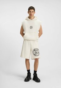 Ung mand iført en ærmeløs cremefarvet hoodie og matchende shorts med boksningsgrafik, sorte sokker og sorte støvler, stående mod en ensfarvet baggrund.