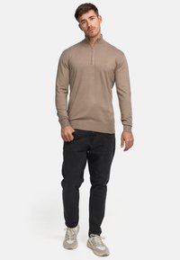 Beige Reißverschluss-Pullover, gerippte Bündchen, schlanker Schnitt, kombiniert mit schwarzen Jeans und grauen Sneakers. Glatte Stricktextur, lässiges Design.