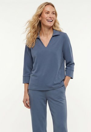 Vrouw met lang blond haar die een bijpassend blauw hemd met kraag en broek draagt, glimlacht met één hand in de broekzak tegen een witte achtergrond.