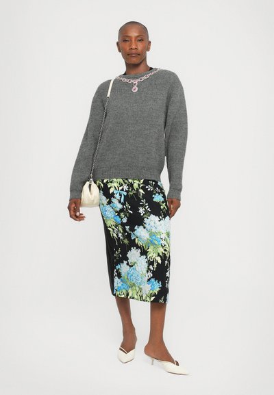 Grauer Strickpullover mit geripptem Muster, schwarzer Rock mit floralen Motiven in Blau und Grün und weißen Kitten-Heel-Schuhen. Kleine weiße Tasche.
