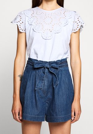 Blouse blanche à manches courtes avec détails en dentelle florale à œillets, associée à un short en denim taille haute avec ceinture nouée à la taille.