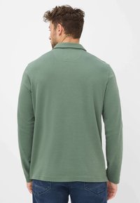 Langärmliges Polohemd aus gedämpftem grünem Stoff. Es hat einen klassischen Kragen, klare Linien und eine lockere Passform. Genossen mit blauen Jeans.