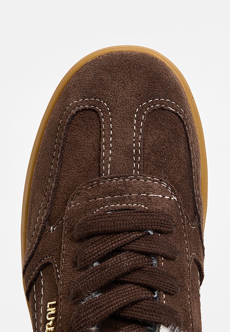 Sneaker in suede marrone con cuciture bianche e lacci marroni, caratterizzato da una tomaia testurizzata e una suola in gomma. Dettaglio del logo dorato sul lato.