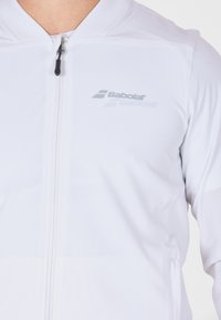 Nærbillede af en hvid zip-up jakke med et synligt Babolat-logo på brystet og en lynlås trækstrop.