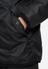 Doudoune noire avec un extérieur lisse et brillant. Comprend une poche zippée visible et des poignets élastiques. Le matériau semble isolant et durable.