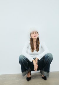 Mladá žena v bielom dlhom rukáve, chlpatou bielou čiapkou, nohaviciach so širokými nohavicami a čiernych špicatých podpätkoch, ktorá sa podopiera o bielu stenu.