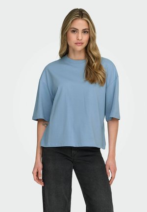 Mujer con cabello largo y rubio, vistiendo una camisa suelta de manga corta azul claro y vaqueros negros, de pie frente a un fondo liso y claro.