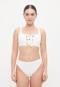 Witte bikini set met een cropped top voorzien van gouden gaten en een vetersluiting, gecombineerd met hoge taille onderkanten. Soepele textuur.