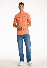 T-shirt med kort ärm i korallfärg med tryck "COPENHAGEN LINDBERGH" på framsidan, tillsammans med blå jeans och vita sneakers. Enfärgad bakgrund.