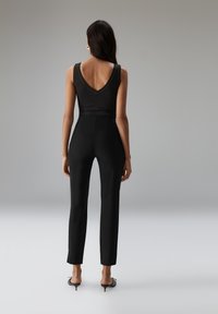 Vrouw met lang donker haar, gekleed in een zwarte mouwloze top met een diepe rug en hooggesneden zwarte broek, staand tegen een effen grijze achtergrond.