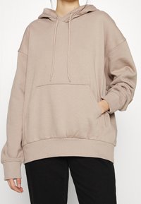 Persona che indossa una felpa oversize beige con tasca frontale e cordini, abbinata a pantaloni neri, su uno sfondo bianco semplice.