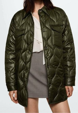 Femme portant une veste matelassée vert foncé sur un chemisier blanc et une jupe pied-de-poule noire et blanche, debout les bras détendus.