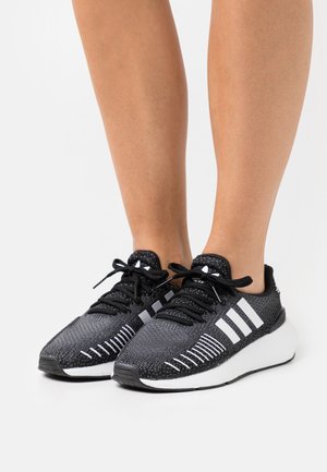 Baskets de sport noires avec une tige en tricot texturé, des rayures blanches, des lacets et une semelle intermédiaire blanche épaisse ; elles présentent un nez arrondi et un talon renforcé.