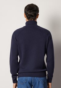 Mörkblå stickad tröja med hög krage, ribbade muddar och fåll, visad bakifrån. Mjuk textur och figursydd design.