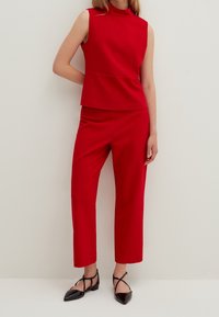 Woman wearing a sleeveless red top with matching red cropped pants and black pointed-toe strappy flats, standing against a white wall.