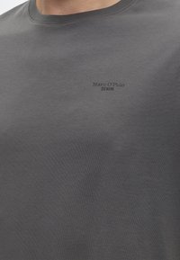 Prim-plan al unui tricou gri cu guler rotund și text mic negru "Marc O'Polo DENIM" pe partea stângă a pieptului.