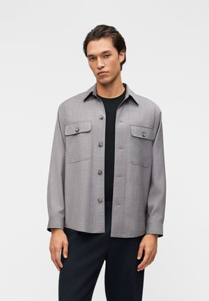 Νεαρός άνδρας που φορά ανοιχτό γκρι πουκάμισο με κουμπιά και τσέπες με καπάκια πάνω από μαύρο πουκάμισο, στέκεται μπροστά σε απλό ανοιχτό φόντο.