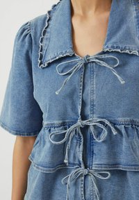 Vaaleansininen farkkumekko, jossa on röyhelökaulus, lyhyet hihat sekä narut vyötäröllä ja rinnassa. Pehmeä rakenne ja kerroksellinen muotoilu.