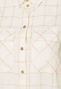 Camicia button-up crema con motivo a griglia dorata, due tasche sul petto e bottoni beige. Il tessuto ha una trama testurizzata e un colletto.