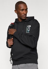 Hoodie preto feito de tecido macio, com um patch de logotipo, capuz com cordão e bolsa canguru. Combinado com calças cinzas e uma mochila.
