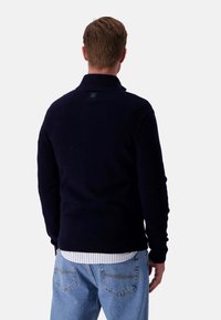 Marineblauer, gestrickter Pullover mit hohem Kragen, gerippten Bündchen und Saum. Über einem weißen, gestreiften Hemd getragen, das ein kleines Logotag auf der Rückseite hat.