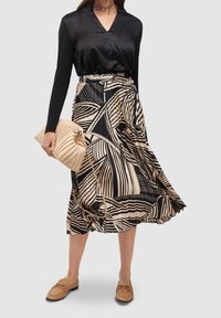 Donna che indossa una blusa nera a maniche lunghe, una gonna midi plissettata con motivo nero e beige, mocassini color sabbia e tiene in mano una borsa a tracolla beige trapuntata.